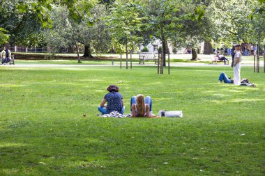 İnsanlar yemyeşil bir şehir parkında piknik battaniyesinde ağaçların altındaki yeşil çimlerde oturup güneş ışığı ve sakin kentsel doğayla sakin bir günün tadını çıkarıyorlar. Londra, İngiltere, 23 Temmuz 2023