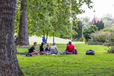 Yeşil bir parkta battaniyelerde dinlenen karışık bir grup, yapraklı ağaçların altında gündelik piknik ve sohbetin keyfini çıkarıyorlar. Londra, İngiltere, 29 Temmuz 2023