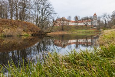 Bir göletin üzerindeki tarihi kale ve malikanenin sakin kırsal manzarası. Ağaçlar ve çimenler yumuşak bulutlu gökyüzünün altında sakin su yansımaları oluşturuyor..
