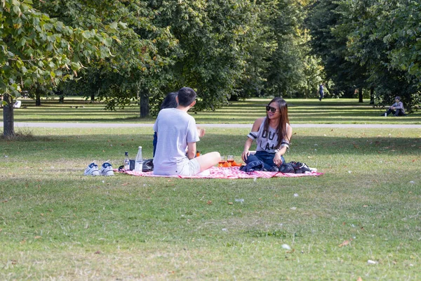 Yeşil bir parkta piknik battaniyesinde oturan çift yapraklı ağaçların altında içki ve atıştırmalıkları paylaşıyor. Rahatlamış açık hava yaşam tarzı, flört, hafta sonu eğlencesi ve şehir doğası. Londra, İngiltere, 28 Ağustos 2023