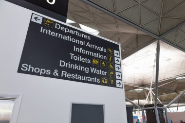 Kalabalık bir havaalanı terminalinde, yolcuları uluslararası geliş, gidiş, tuvalet, içme suyu ve dükkanlara yönlendiren açık, siyah bir bilgi işareti. Londra, İngiltere, 18 Kasım 2023