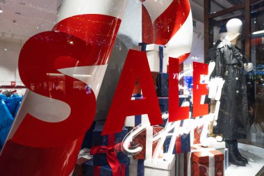 Cam dükkanının penceresinde büyük bir kırmızı satış tabelası var. İçinde bayram süslemeleri, hediye kutuları ve arka planda ceket giyen bir manken var..