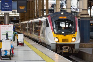 Yoğun bir şehir tren istasyonunda kalkışa hazır modern elektrikli yolcu treni bekliyor. Arka planda banliyö sakinleri ve dijital tabelalar görülüyor. Londra, İngiltere, 11 Şubat 2024