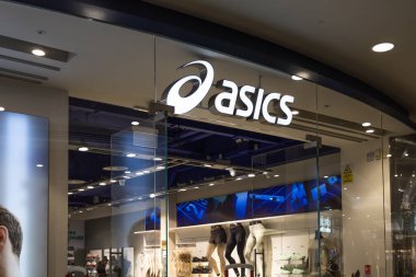 Modern bir alışveriş merkezi ortamında cam bir vitrinin üzerinde aydınlatılmış beyaz logo ve spiral ikonun yer aldığı bir Asics mağazası girişinin görüntüsü. Londra, İngiltere, 17 Şubat 2024