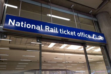 İngiliz tren istasyonunun cam girişinde asılı mavi bir Ulusal Demiryolu bilet ofisi tabelası, toplu taşımayı ve İngiliz demiryolu altyapısını temsil ediyor..