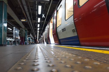 Londra metro istasyonu peronunun düşük açılı görüntüsü. Görme engelliler için sarı dokunsal kaldırımlar ve açık kapıları olan bir tren var. Londra, İngiltere, 3 Mart 2024