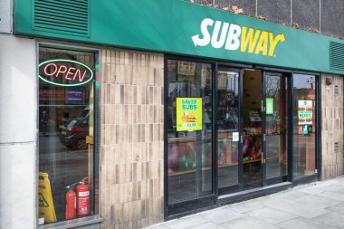 Metro sandviç dükkanının sokak manzaralı bir mağazası. Girişte cam kapılar, tanıtım posterleri ve tuğla bir cephede neon açık tabela yer alıyor. Londra, İngiltere, 3 Mart 2024