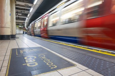 Londra metro istasyonunda kırmızı ve beyaz bir banliyö treninin yüksek hızlı hareket bulanıklığı. Ön planda Gap güvenlik levhası var..