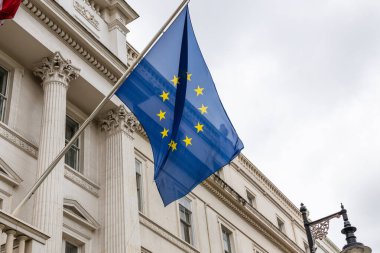 Üzerinde sarı yıldızlar bulunan mavi bir Avrupa Birliği bayrağı bulutlu bir gökyüzünün altında sütunları olan beyaz bir klasik binaya bağlı bir bayrak direğinden dalgalanır..