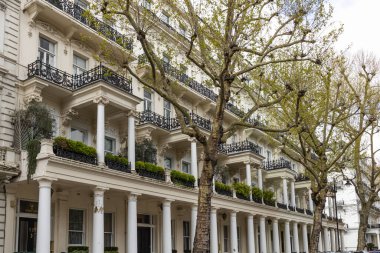 Klasik sütunları ve siyah demir balkonları olan zarif beyaz sıva evlerinin alçak açılı görüntüsü. Lüks konutların önündeki caddede yeşil ağaçlar var..