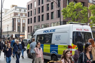 Londra 'da kalabalık bir kaldırımda konuşlanmış, kalabalık yayalar ve şehir binalarıyla çevrili beyaz ve sarı bir polis minibüsü. Londra, İngiltere, 14 Nisan 2024