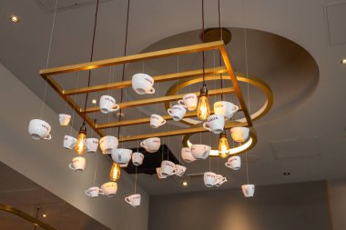 Beyaz Costa Kahve fincanlarından yapılmış eşsiz bir avizenin şık bir iç çekimi ve altın çerçeveden sarkan sıcak klasik ampuller. Londra, İngiltere, 27 Mayıs 2024