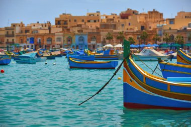 Malta 'nın renkli adası. Turkuaz denizde turuncu antik binalara karşı mavi-sarı balıkçı tekneleri. Balıkçı limanı, su yüzeyinden manzara. Malta 'da tatiller.