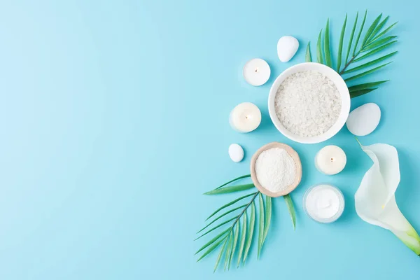 Spa background with skin care products, stones, candles and palm leaves on blue. Flat lay, copy space