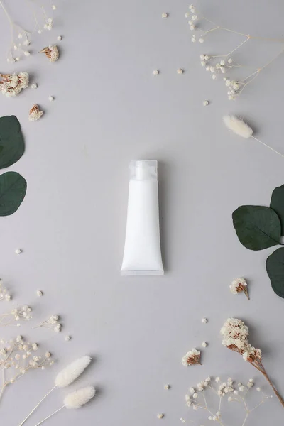 White tube of cosmetic cream with flowers and green leaves on grey background. Flat lay