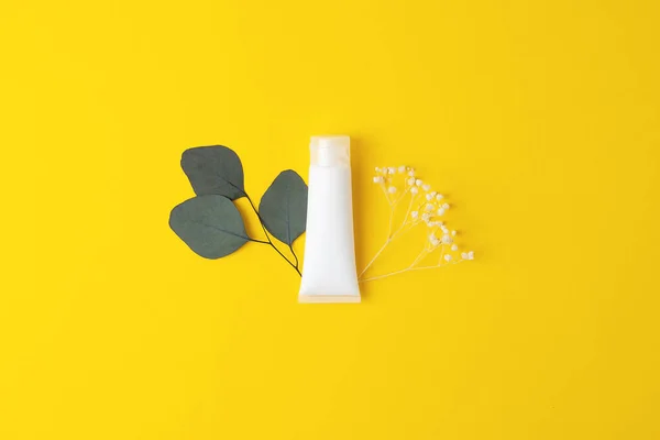 White tube of cosmetic cream with flowers and green leaves on yellow background. Flat lay, copy space