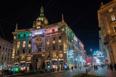 Milan, İtalya, 11 Şubat 2026: İtalyan bayrağının renkleriyle aydınlatılmış zarif tarihi bir binayla Merkez Milano. Dekoratif ışıklar tramvayların üzerindeki caddeyi kaplarken, yayalar dükkanlar arasında dolaşıyorlar..