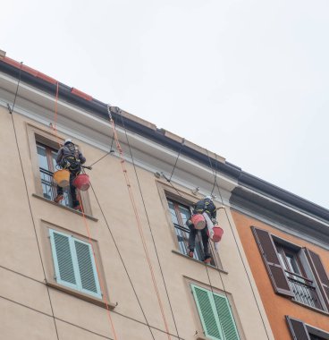 Milan İtalya 27 Şubat 2026: Bir binanın ön cephesinde askıya alınmış, güvenlik ekipmanları ve teçhizatlarıyla yüksek bakım ve yenileme çalışmaları yapan iki uzman işçi bir halat üzerinde çalışıyor.