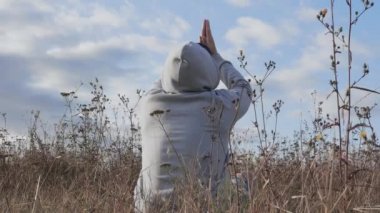 Doğada meditasyon yapan, gökyüzüne ve bulutlara bakan kapüşonlu ve kapüşonlu bir adam yoga egzersizleri yapıyor.
