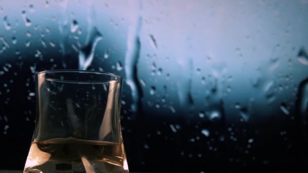 一杯酒在雨的背景下 威士忌在玻璃中 雨水在窗外 水从玻璃杯中流下来 美丽的水滴 — 图库视频影像
