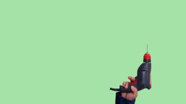 Electric drill in hand on a green background, construction tool. A man in construction overalls holds a drill in his hand.