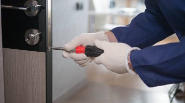 The interior door is being repaired by a handyman, a man in a special uniform with a screwdriver in his hands, a carpenter at work