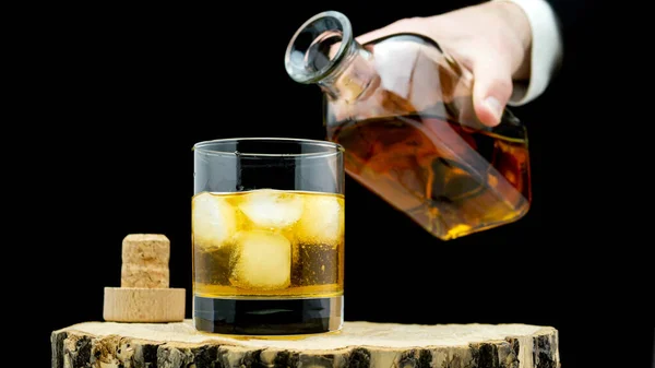 Pour whiskey into a glass with ice on a rustic on a black background. Whiskey in a clear glass