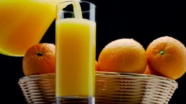 Siyah arka plandaki bir bardağa taze ananas suyu dök. Tahtadaki saman sepetinde portakallar.