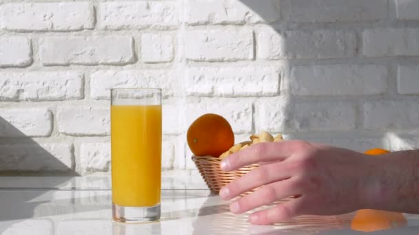 男は手に黄色いレモネードを持っている 白いレンガの壁を背景に白いテーブルの上にガラスのオレンジジュース — ストック動画