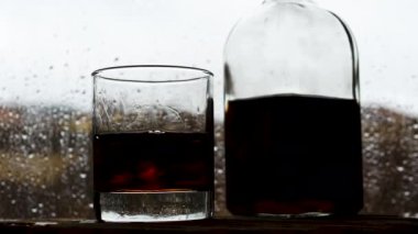 Yağmur penceresinin yanındaki barın üstünde bir şişe viski. Damlalar camdan aşağı akıyor. Barda alkol..