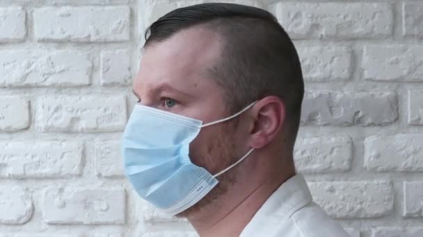 白いレンガの壁を背景に 彼の顔に医療用マスクのハンサムな男性医師の肖像画 その男は顔を医療マスクで覆った 幸せな男はウイルスから保護されています 健康コンセプト — ストック動画