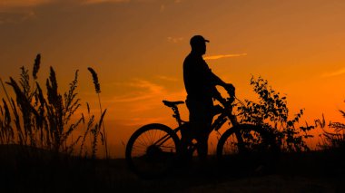 Gün batımında bisikletli bir siluet adam. Bisikletçi güzel bir turuncu gün batımına bakıyor. Dağın zirvesinde yaşayan bir şampiyon.