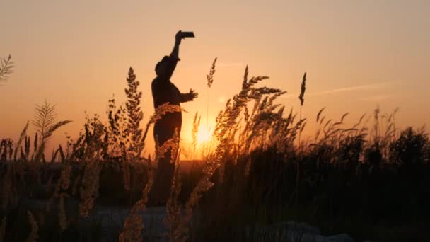 最快乐的人一个拿着智能手机的快乐男人拿着一个自拍照的轮廓 一个站在美丽日落的背景下的男人 — 图库视频影像