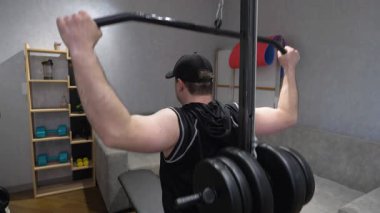 Genç ve yakışıklı sporcu sırt kaslarını ağır bir simülatörle şişiriyor. Modern apartmanlarda evde antrenman. Yavaş çekim