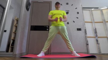 Atletik genç adam elinde dambıllarla omuzlarını pompalıyor. Evde spor yaparken, bir apartman dairesinde spor yaparken. Yavaş çekim.