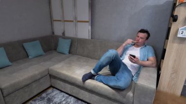 Dişleri diş fırçasıyla fırçalayan bir adam oturma odasında kanepede oturuyor. Dairesinde diş macunuyla diş fırçalamak. Yavaş çekim.