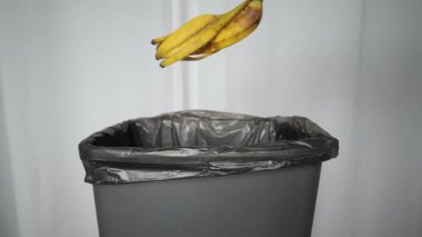 Bir adamın eli muz kabuğunu yavaş çekimde çöp kutusuna atar..