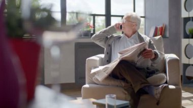 yaşlı adam bir sandalyede oturur, gazete okuma ve modern bir dairede telefonda konuşmak