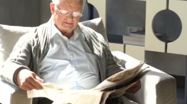 mutlu yaşlı adam bir sandalyede oturur ve modern bir dairede bir gazete okuma