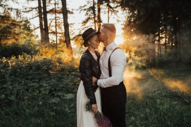 Günbatımında ormanda öpüşen hippi düğün çifti.