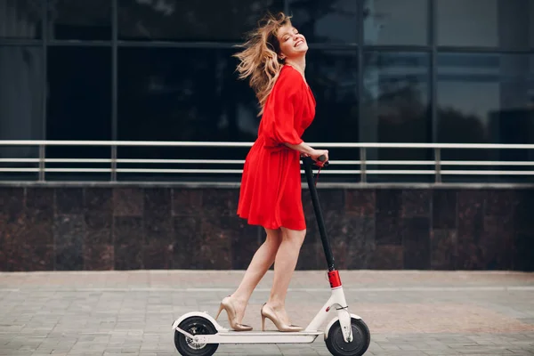 Young woman riding electric scooter in red dress and high-heeled shoes