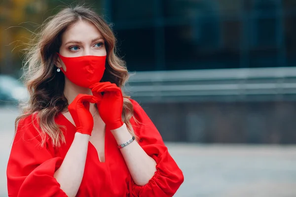 Jonge vrouw portret in rode jurk en handschoenen zet op medisch gezichtsmasker op straat buiten — Stockfoto