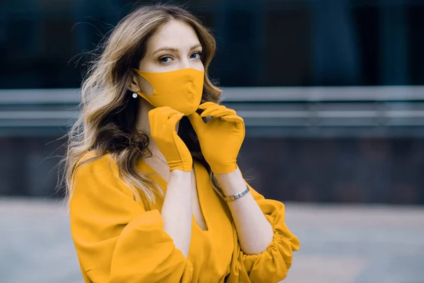 Junge Frau in gelbem Kleid und Handschuhen setzt auf der Straße einen medizinischen Mundschutz auf. Coronavirus covid-19 Quarantäne-Konzept. — Stockfoto