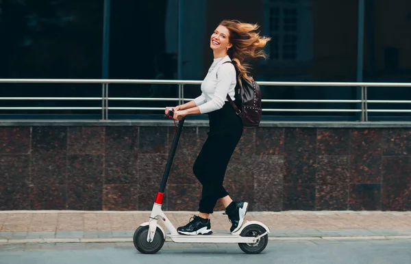 Wanita muda dengan senyum naik skuter listrik di jalan Stok Lukisan  