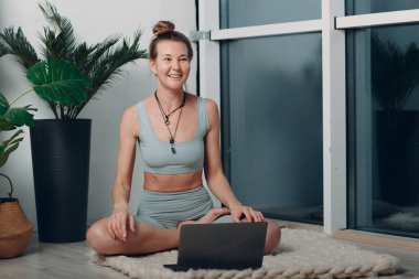 Yetişkin, gülümseyen bir kadın evinde yoga yapıyor. Bilgisayarında online dersler var.