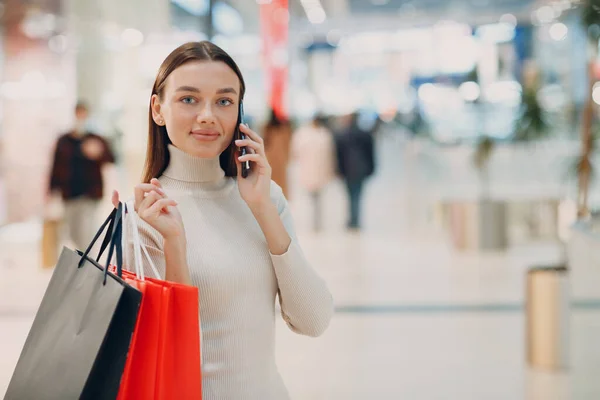 Mujer adulta joven positiva que lleva bolsas de papel y teléfono móvil en las manos —  Fotos de Stock