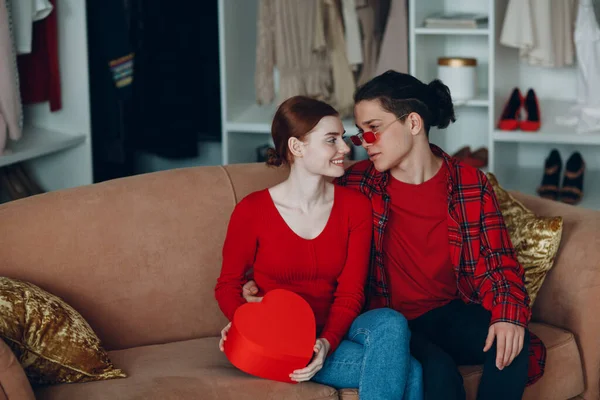 Sorrindo jovem mulher e homem com namorados caixa de presente coração vermelho em forma de sofá em casa — Fotografia de Stock