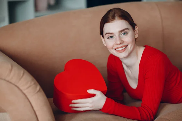 Cantik wanita muda dengan rambut merah tergeletak di sofa di ruang tamu dan memegang kotak hadiah dalam bentuk hati dan tersenyum. Hari Valentine atau Ulang Tahun — Stok Foto