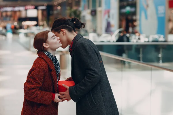 Ein Kerl schenkt seiner Freundin zum Valentinstag eine herzförmige Schachtel. Junges Paar küsst sich in Einkaufszentrum. — Stockfoto