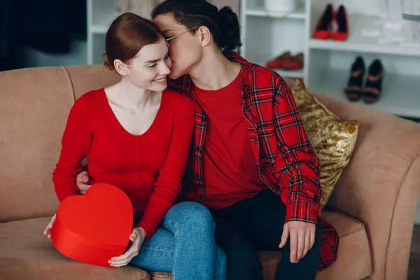 Lächelnde junge Frau und Mann mit Valentinstag Geschenkbox rotes Herz geformt zu Hause Sofa — Stockfoto